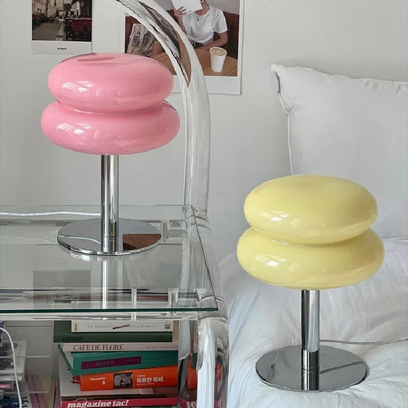 Two colorful stools, one pink and one yellow, on a glass shelf with books and a white couch in the background.