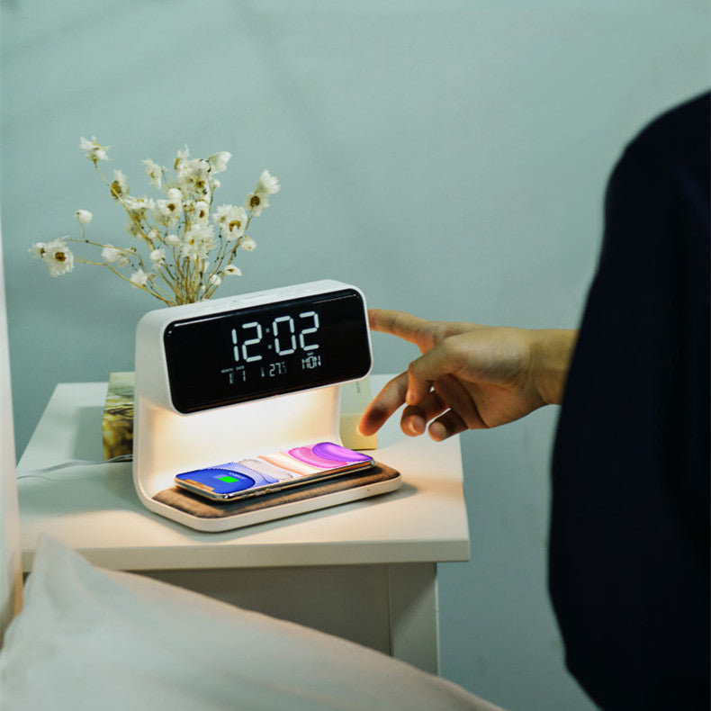 Digital clock with phone charging station on a nightstand, with a hand interacting with it.