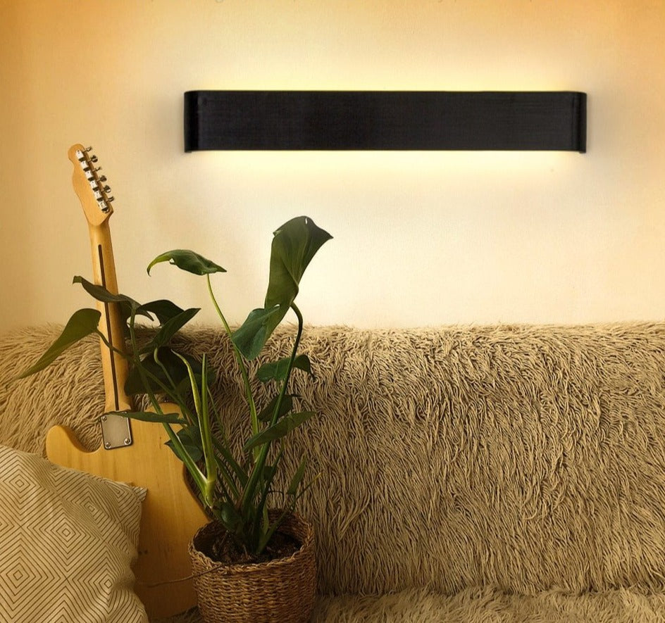 Guitar leaning against a wall with a potted plant on a couch, illuminated by a wall-mounted light fixture.