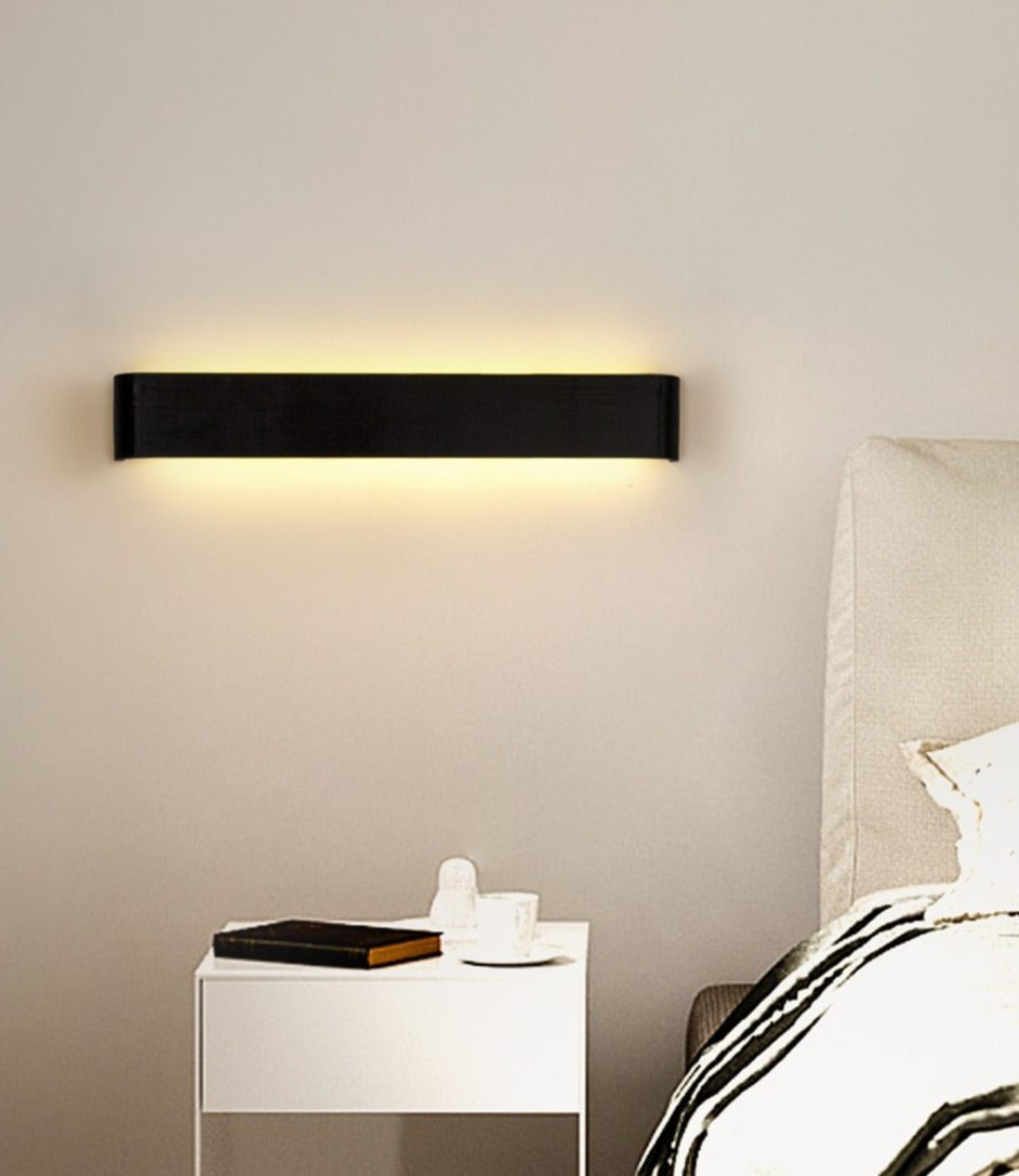 Modern wall light fixture on a neutral wall above a nightstand with a book and small round object.