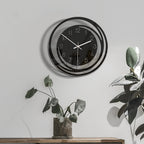 Modern wall clock with black and silver design on a white wall with plants.