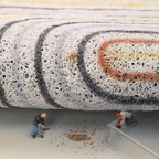 Model of a large roll of carpet with two small figures working on it.