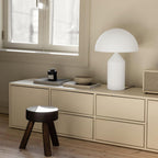 Modern interior with a white table lamp, wooden stool, and decorative items on a beige cabinet.