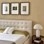 Bedroom with a tufted headboard, framed pictures on the wall, and a side table with a lamp.