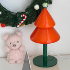 Orange Christmas tree-shaped lamp with a green base next to a pink teddy bear and festive decorations.