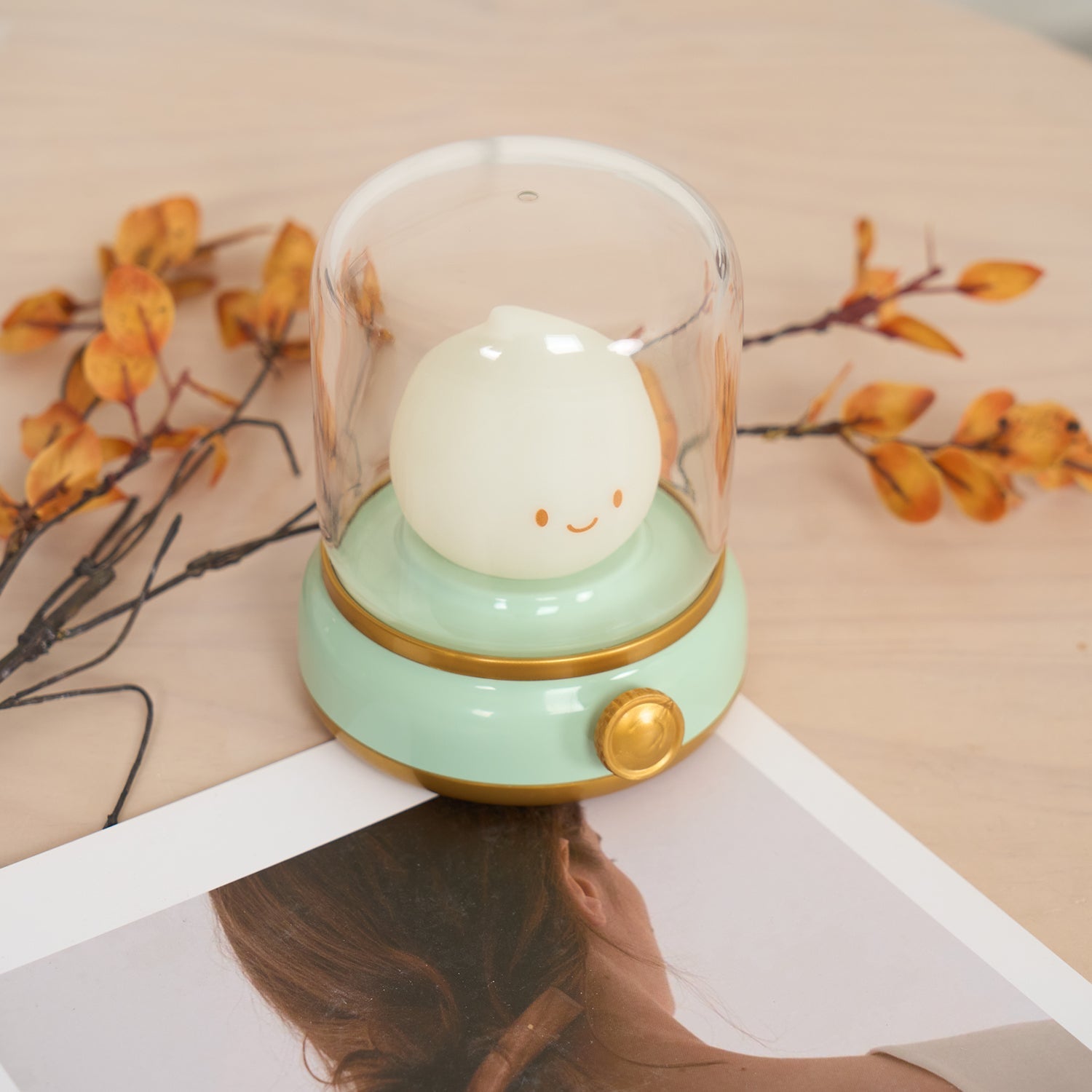 Decorative lamp with a smiling face inside a glass dome on a surface with autumn leaves.
